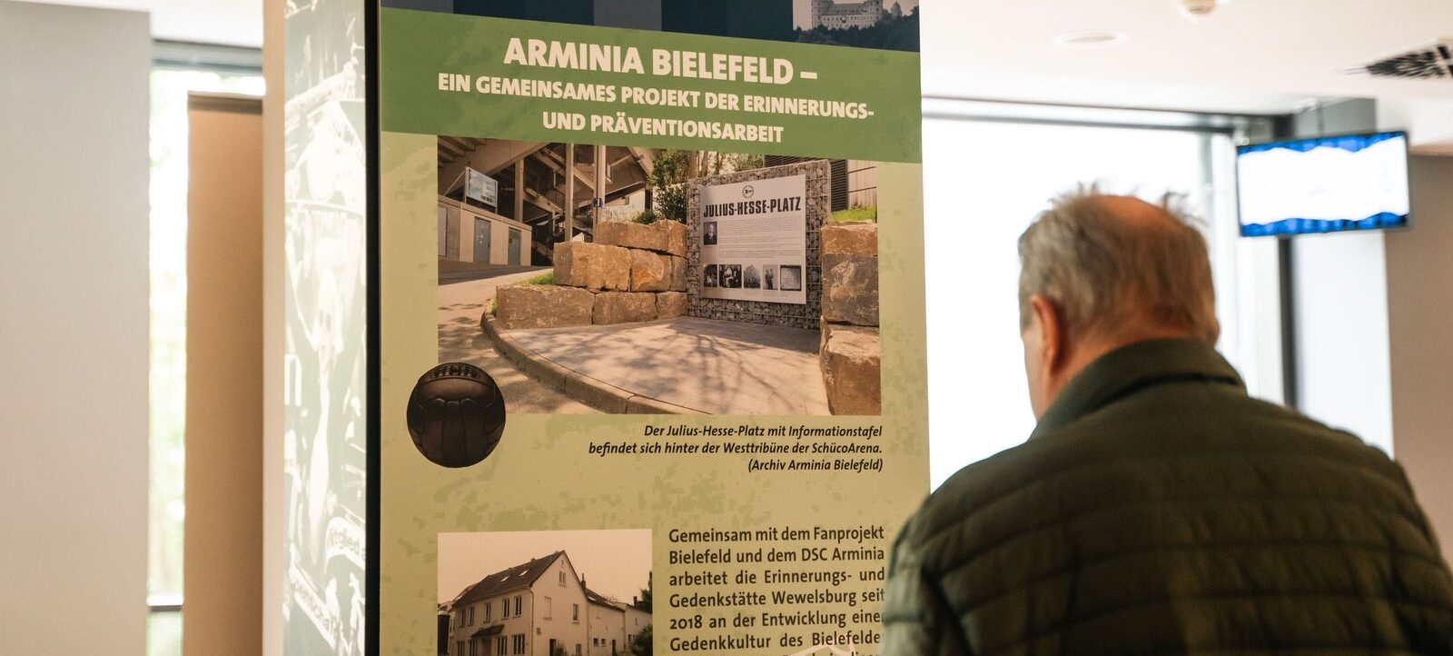 Ein Mann betrachtet eine Informationsplakat zum Projekt "Arminia Bielefeld – Ein gemeinsames Projekt der Erinnerungs- und Präventionsarbeit". Das Plakat zeigt Fotos des Julius-Hesse-Platzes mit Informationstafel und eines ehemaligen Vereinslokals.