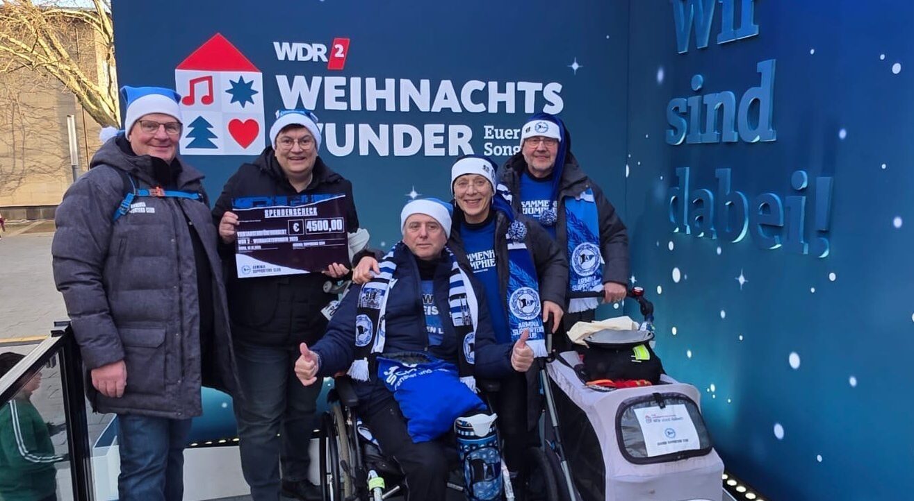 Eine Gruppe von fünf Personen, darunter eine im Rollstuhl, steht vor einer blauen Wand mit der Aufschrift "WDR 2 Weihnachtswunder". Die Personen tragen blaue Mützen und Schals mit einem Logo und lächeln in die Kamera. Eine Person hält einen Scheck über 4500 Euro in den Händen. Im Hintergrund ist ein winterlicher Himmel mit Schneeflocken zu sehen.