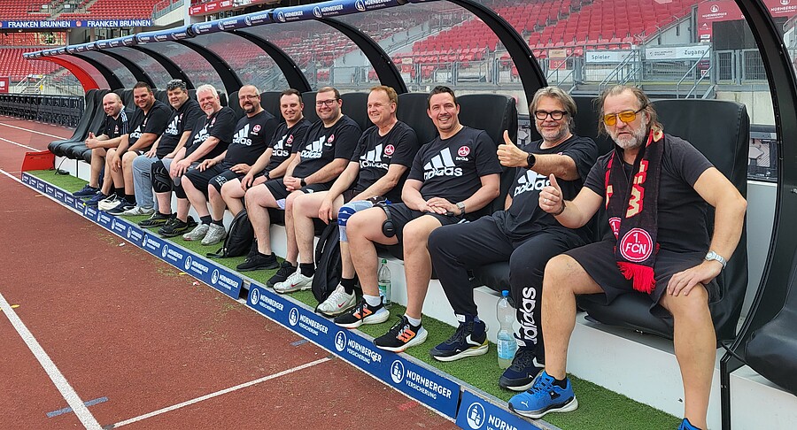 Eine Gruppe von Männern sitzt in einer Stadionbank und lächelt in die Kamera. Sie tragen schwarze Sportkleidung mit dem Logo des 1. FC Nürnberg. Der Hintergrund zeigt einen Teil des Stadions mit roten Sitzen.