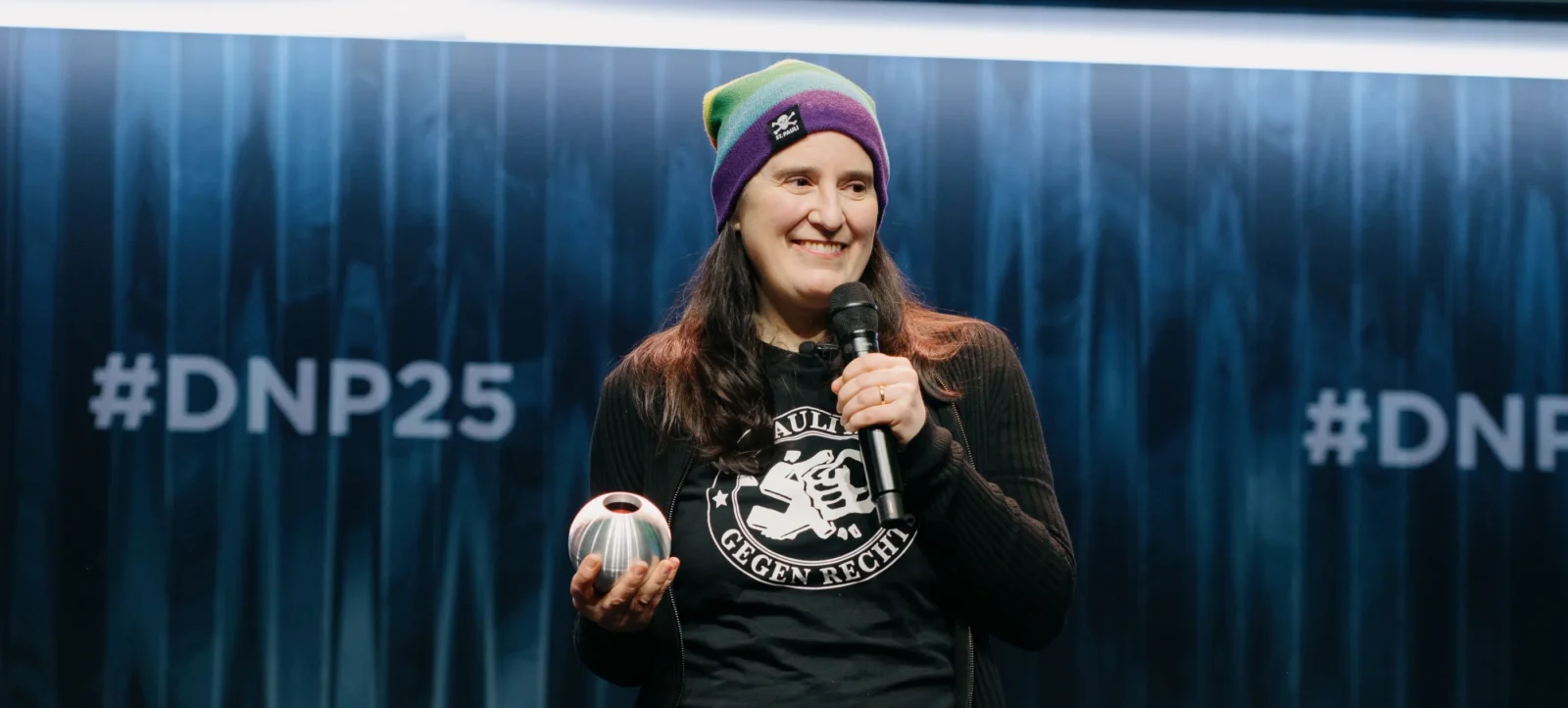 Eine lächelnde Person mit einer bunten Mütze und einem schwarzen T-Shirt mit einem Aufdruck hält ein Mikrofon in der einen Hand und einen metallischen Gegenstand in der anderen. Im Hintergrund ist die Aufschrift "#DNP25" zu sehen.