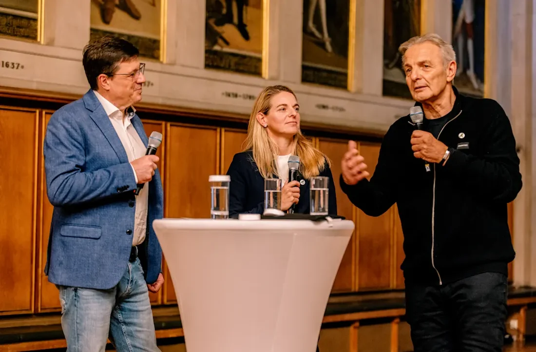 Drei Personen stehen hinter einem Pult und halten Mikrofone in den Händen. Links ein Mann im Anzug, in der Mitte eine Frau mit blonden Haaren und rechts ein älterer Mann in einem schwarzen Sweatshirt. Sie scheinen in einer Diskussion oder einem Gespräch auf einer Bühne zu sein. Im Hintergrund sind Holzvertäfelungen und Bilder zu sehen.