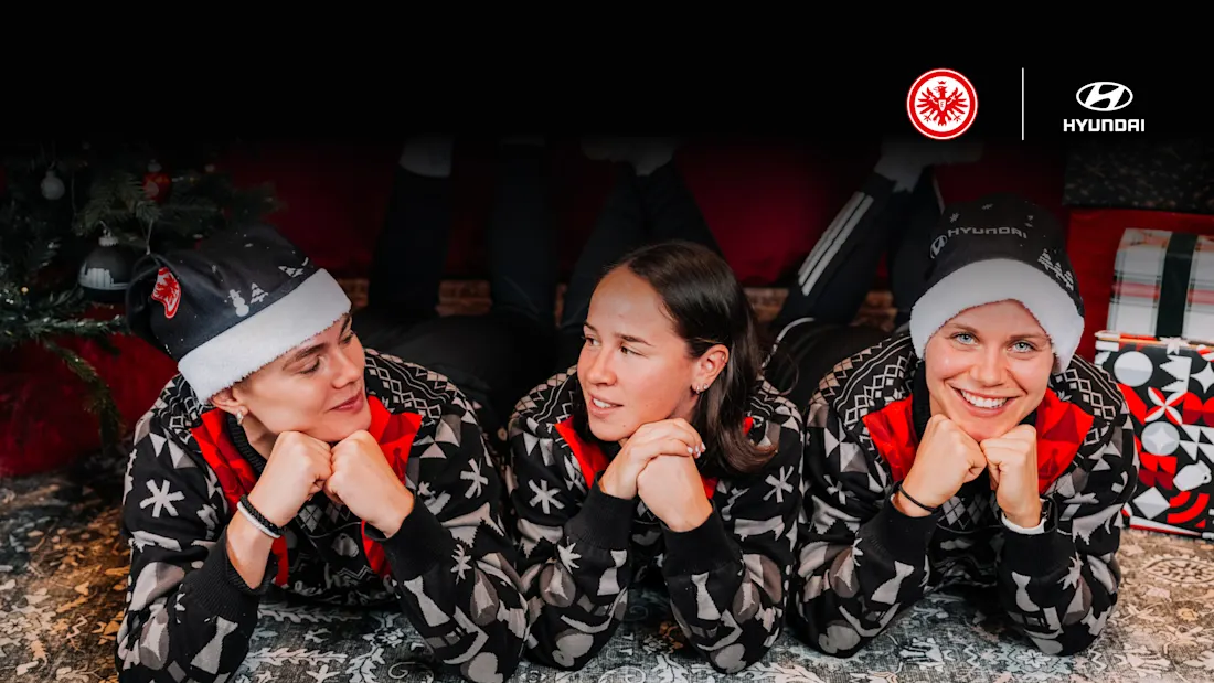 Drei Personen in Weihnachtsmützen und passenden Pyjamas liegen auf dem Boden und lächeln in die Kamera. Im Hintergrund sind Weihnachtsdekorationen und Geschenke zu sehen. Die Logos von Eintracht Frankfurt und Hyundai sind im oberen rechten Eck zu sehen.