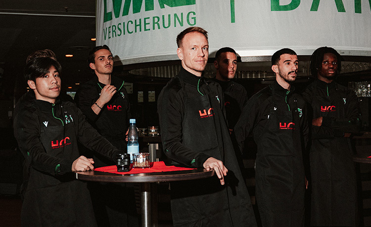 Eine Gruppe von Menschen in schwarzer Kleidung mit grünen Akzenten und dem Logo "HCC" steht hinter einem Tisch. Auf dem Tisch befinden sich eine Wasserflasche und einige Gläser. Im Hintergrund ist ein Banner mit der Aufschrift "VERSICHERUNG" zu sehen. Die Personen scheinen Teil eines Teams zu sein und sind möglicherweise bei einem Event oder einer Präsentation.