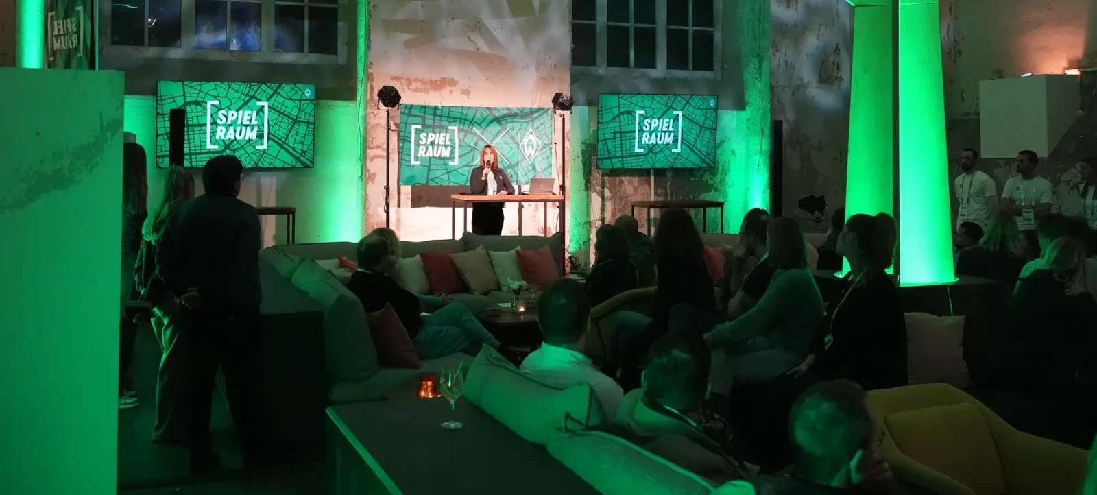 Eine Frau steht an einem Pult und spricht in einen Mikrofon. Sie ist von einem grünen Licht beleuchtet und befindet sich in einem Raum mit dem Namen "Spielraum". Der Raum ist mit bequemen Sitzgelegenheiten ausgestattet, auf denen mehrere Personen sitzen und der Frau zuhören. Im Hintergrund sind weitere Personen zu sehen, die sich unterhalten oder dem Vortrag folgen. Die Wände des Raumes sind mit Graffiti verziert und es gibt große Fenster, durch die Tageslicht hereinfällt.