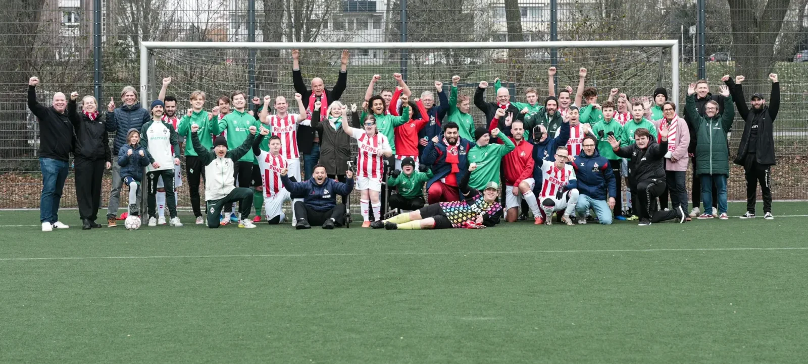 Eine Gruppe von Menschen steht vor einem Fußballtor auf einem grünen Kunstrasenfeld. Die Personen tragen verschiedene Trikots und Kleidung, darunter grüne, rote und weiße Farben. Einige Personen sind in die Hocke gegangen, während andere stehen. Alle lächeln und heben die Fäuste in die Luft, was auf eine erfolgreiche oder freudige Veranstaltung hindeutet. Im Hintergrund sind ein Zaun und Bäume zu sehen.