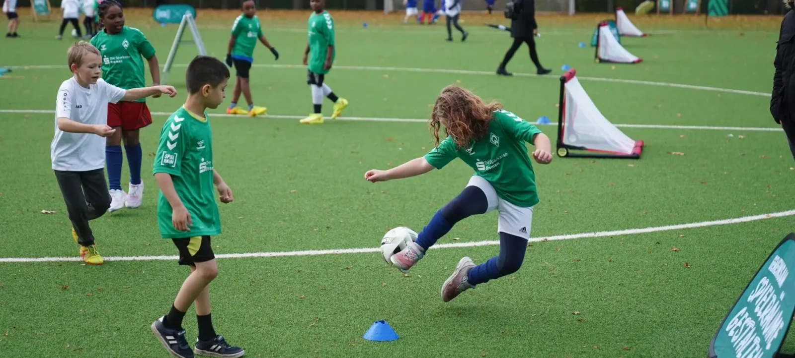 Kinder in grünen und weißen Trikots spielen auf einem Fußballfeld. Ein Mädchen schießt einen Fußball durch ein Mini-Tor, während andere Kinder in der Nähe stehen und zuschauen. Im Hintergrund sind weitere Kinder und ein Tor zu sehen.