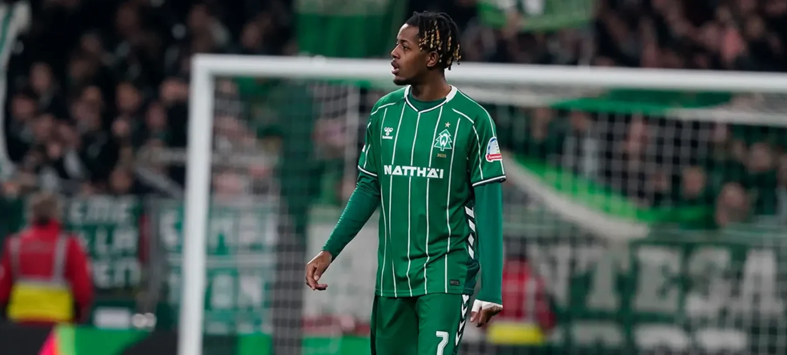 Ein Fußballspieler im grünen Trikot mit der Nummer 7 steht auf dem Spielfeld vor dem Tor. Im Hintergrund sind Zuschauer und ein Teil des Stadions zu sehen.