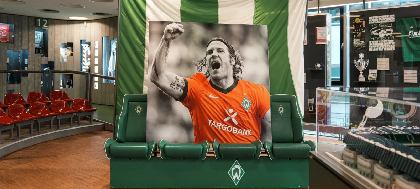 Ein Spieler des Vereins Werder Bremen in einem orangefarbenen Trikot mit dem Logo von Targobank posiert vor einem grün-weißen Hintergrund, der an die Vereinsfarben erinnert. Er sitzt auf einem grünen Sitzplatz, der mit dem Vereinswappen verziert ist, und hebt triumphierend die Faust. Im Hintergrund sind rote Sitzplätze und eine Vitrine mit Trophäen und Erinnerungsstücken zu sehen.