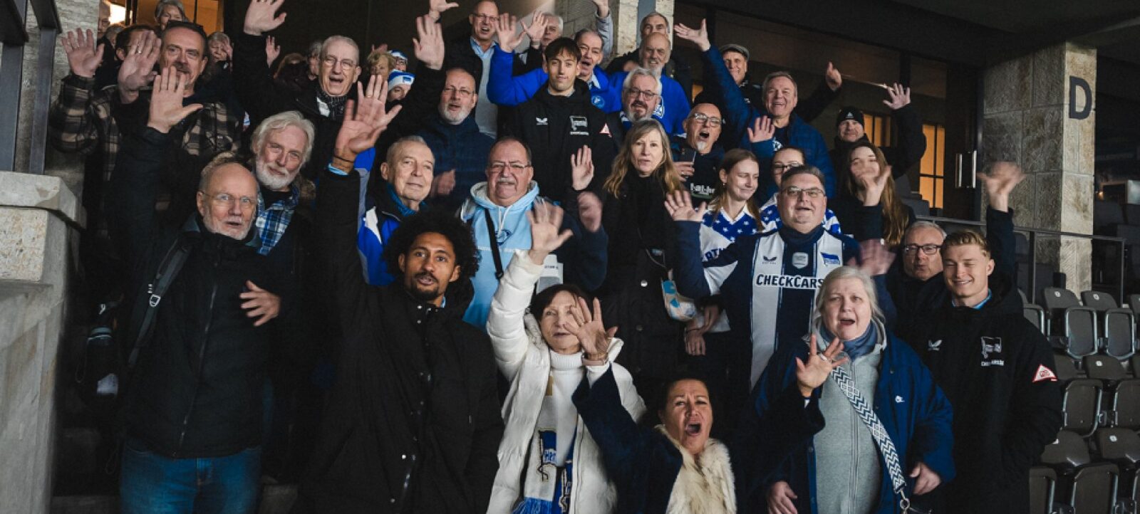 Eine Gruppe von Menschen, darunter Fans in blau-weißen Schals und Trikots, posieren fröhlich für ein Gruppenfoto. Sie stehen auf einer Treppe vor einem Gebäude und winken oder machen lustige Gesten. Die Stimmung ist ausgelassen und freundschaftlich.