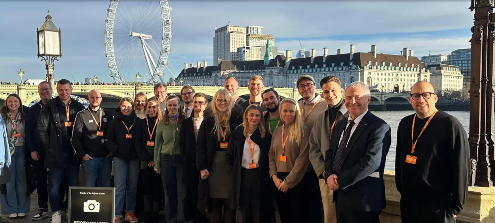 Eine Gruppe von Menschen steht für ein Gruppenfoto am Ufer der Themse in London. Im Hintergrund sind das London Eye und historische Gebäude zu sehen. Die Personen tragen verschiedene Kleidungsstücke und einige haben Namensschilder um den Hals. Die Atmosphäre wirkt freundlich und professionell.