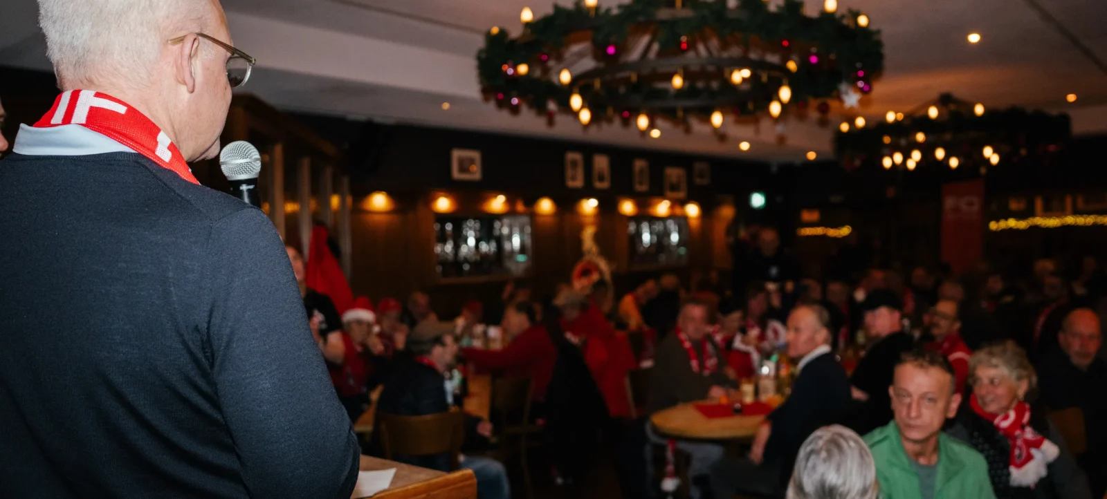 Ein Mann mit weißem Haar und einer roten und weißen Schärpe spricht in ein Mikrofon vor einer Gruppe von Menschen in einem festlich dekorierten Raum. Der Raum ist mit Lichtern und einem Kranz geschmückt, und die Menschen tragen festliche Kleidung, darunter Weihnachtsmützen und Schals.