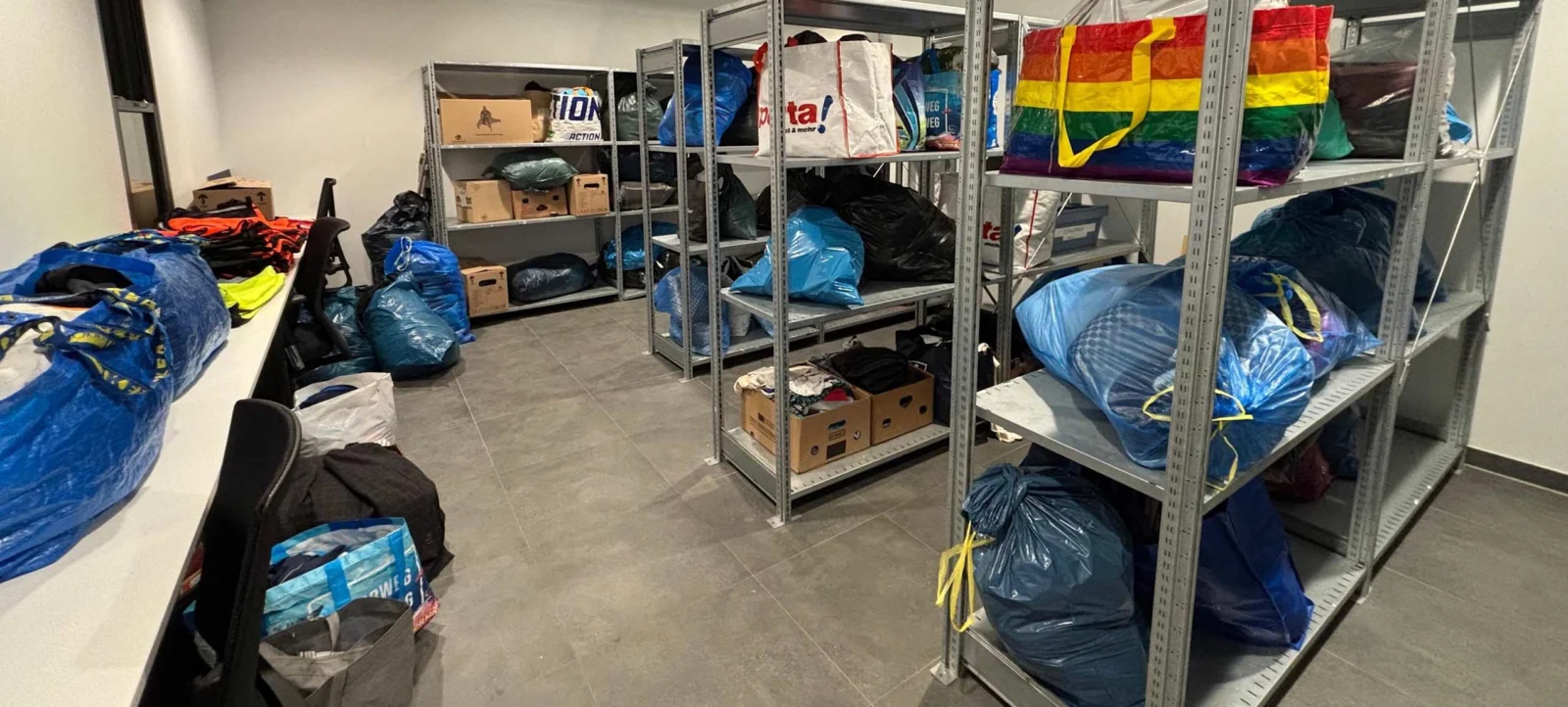 Ein Lagerraum mit vollen Regalen und Säcken voller Kleidung. Die Säcke sind in verschiedenen Farben, darunter blau, schwarz und Regenbogenfarben. Der Raum ist mit Kisten und weiteren Kleidungsstücken auf dem Boden gefüllt.
