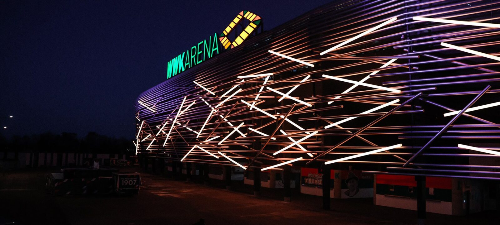 Nachtszene vor der WWK ARENA mit Lichtstreifen und leuchtendem Schriftzug. Die Lichtstreifen erzeugen dynamische Muster auf der Fassade des Stadions, was eine lebendige und moderne Atmosphäre vermittelt.