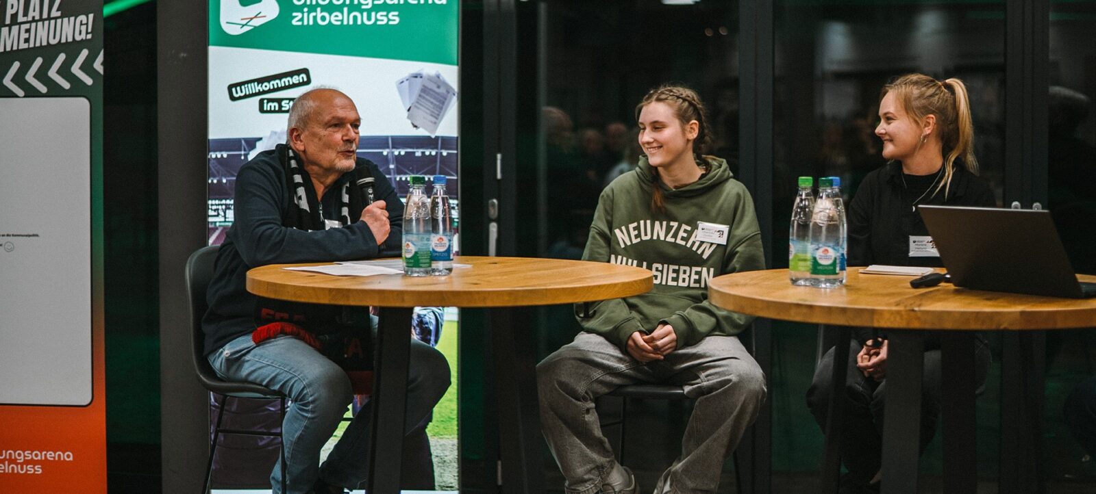 Eine Diskussionsrunde in der Bildungsarena Zirbelnuss. Ein älterer Mann sitzt links und spricht, während zwei junge Frauen rechts zuhören und Notizen machen. Auf dem Tisch stehen Wasserflaschen und Laptops. Im Hintergrund ist ein Banner mit dem Logo der Bildungsarena Zirbelnuss und der Aufschrift 'Willkommen im Stadion' zu sehen.