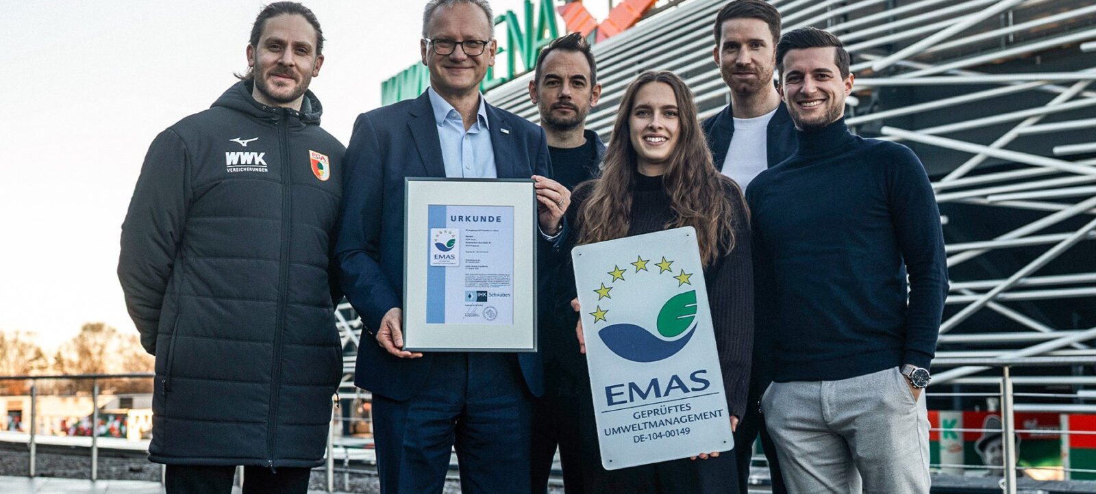 Eine Gruppe von sechs Personen steht vor einem Stadion und hält zwei Urkunden. Die Urkunden tragen die Aufschrift "EMAS" und "Geprüftes Umweltmanagement". Die Personen lächeln und scheinen sich über einen Erfolg im Bereich Umweltmanagement zu freuen.