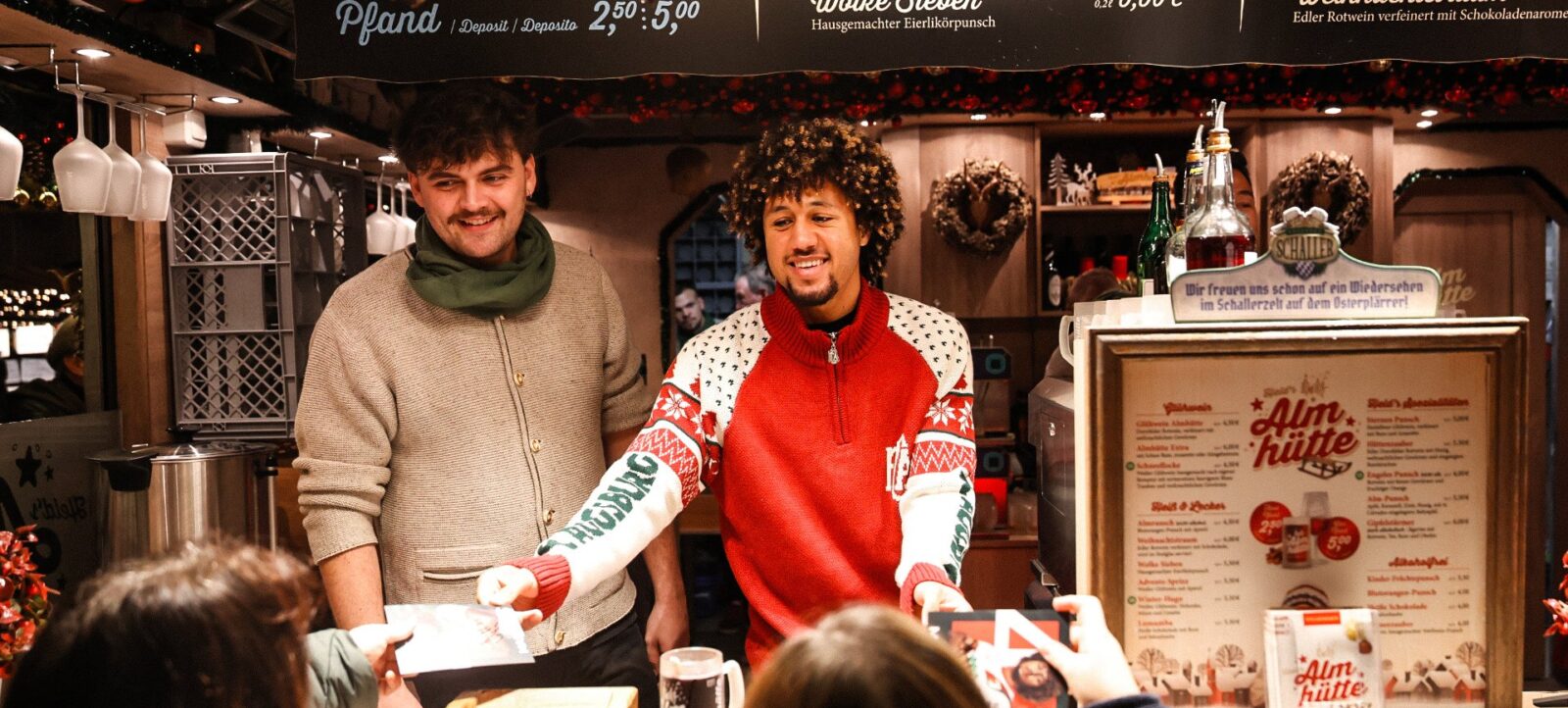 Zwei Personen stehen hinter einem Verkaufsstand auf einem Weihnachtsmarkt. Die Person links trägt einen grünen Schal und einen beigen Pullover, während die Person rechts einen roten Pullover mit weißen Mustern und einen weißen Schal trägt. Beide lächeln und scheinen sich zu amüsieren. Der Stand ist mit weihnachtlichen Dekorationen geschmückt, und es gibt verschiedene Getränke und Speisen auf der Theke. Im Hintergrund sind Preistafeln und ein Schild mit der Aufschrift "Wir freuen uns schon auf ein Wiedersehen im Schallerzelt auf dem Ostermärkt!" zu sehen.