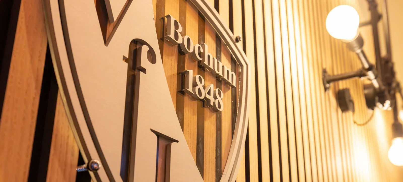 Ein Detailbild des Logos des Vereins VfL Bochum, das ein stilisiertes 'V' und die Aufschrift 'Bochum 1848' zeigt. Das Logo ist an einer Holzwand mit vertikalen Streifen befestigt und wird von einer Glühbirnen-Lampe beleuchtet.
