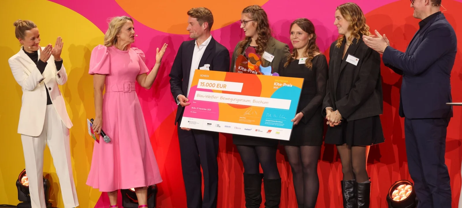 Eine Gruppe von Menschen auf einer Bühne, die einen Scheck über 15.000 Euro für das Projekt "Blau-Weißer Bewegungsraum Bochum" überreichen. Die Personen sind lächelnd und klatschen, während sie vor einem farbenfrohen Hintergrund stehen.