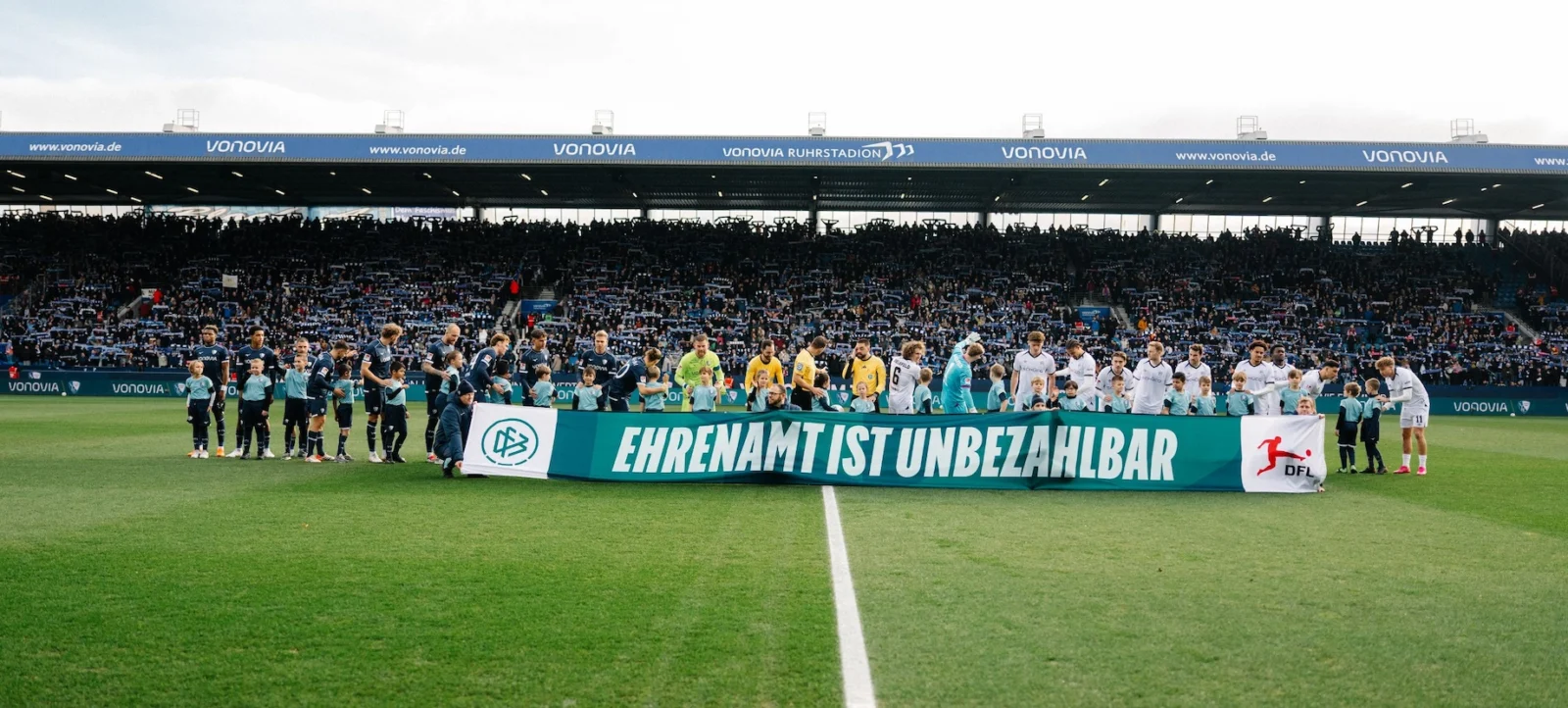 Ein Bild von einem Fußballstadion, auf dem zwei Mannschaften und eine Gruppe von Ehrenamtlichen vor einem Spiel auf dem Rasen stehen. Die Ehrenamtlichen halten ein großes Banner mit der Aufschrift "Ehrenamt ist unbezahlbar" und dem Logo der DFL. Die Tribünen im Hintergrund sind mit Zuschauern gefüllt.