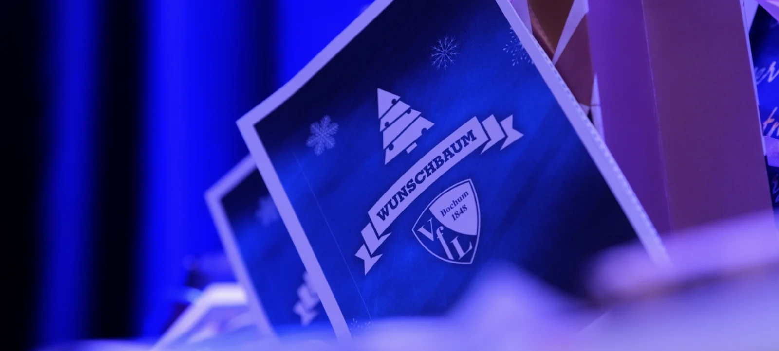 Eine Nahaufnahme eines Weihnachtsprojekts des VfL Bochum. Im Fokus steht eine blaue Karte mit dem Text 'WUNSCHBAUM' und dem Logo des VfL Bochum. Im Hintergrund sind weitere ähnliche Karten zu sehen, die alle mit Schneeflocken und einem Tannenbaum verziert sind.