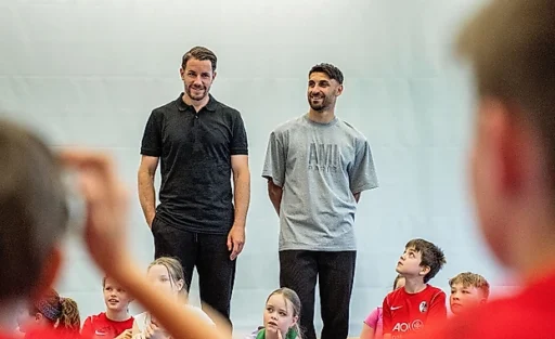 Zwei Männer stehen vor einer Gruppe von Kindern in einer Sporthalle. Die Männer scheinen eine Art Präsentation oder Unterricht zu halten, während die Kinder aufmerksam zuhören. Die Kinder tragen Sportkleidung, darunter T-Shirts mit dem Logo "A07". Die Männer lächeln und wirken entspannt, während sie mit den Kindern interagieren.