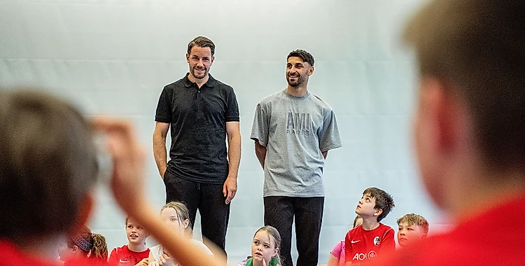Zwei Männer stehen vor einer Gruppe von Kindern in einer Sporthalle. Die Männer scheinen eine Art Präsentation oder Unterricht zu halten, während die Kinder aufmerksam zuhören. Die Kinder tragen Sportkleidung, darunter T-Shirts mit dem Logo "A07". Die Männer lächeln und wirken entspannt, während sie mit den Kindern interagieren.