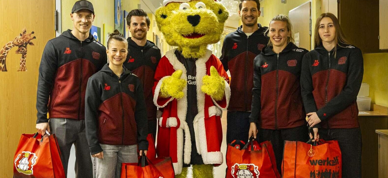 Eine Gruppe von sechs Personen in schwarzen und roten Jacken steht lächelnd neben einem großen, gelben Maskottchen, das wie der Weihnachtsmann gekleidet ist. Sie halten rote Einkaufstaschen mit dem Logo von Bayer 04 Leverkusen in den Händen. Im Hintergrund ist ein Giraffen-Poster an der Wand zu sehen.