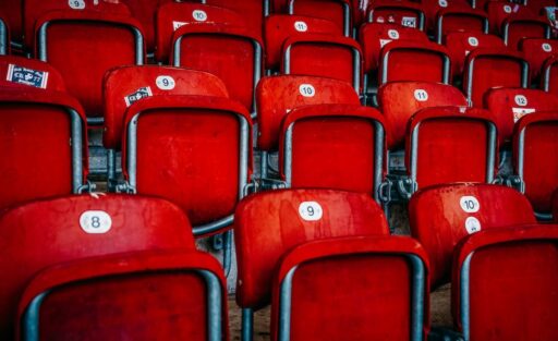 Rote Sitzplätze in einem Stadion, nummeriert und in Reihen angeordnet. Die Sitze sind aus Kunststoff und haben metallische Armlehnen. Die Nummern auf den Sitzen reichen von 8 bis 12.