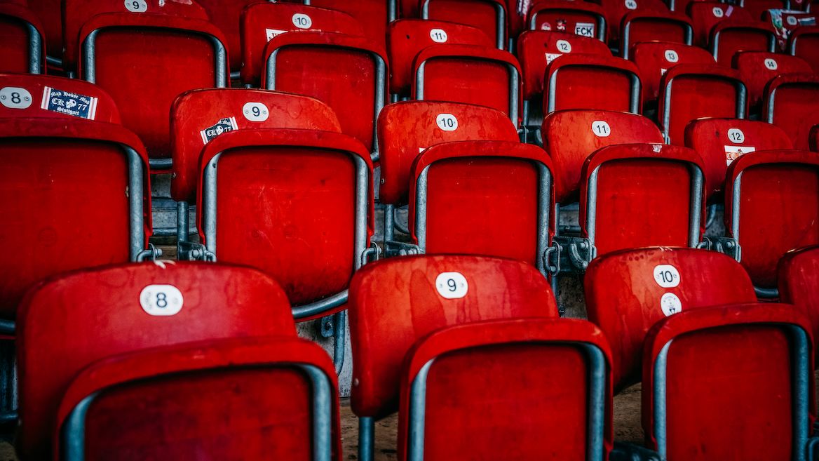 Rote Sitzplätze in einem Stadion, nummeriert und in Reihen angeordnet. Die Sitze sind aus Kunststoff und haben metallische Armlehnen. Die Nummern auf den Sitzen reichen von 8 bis 12.