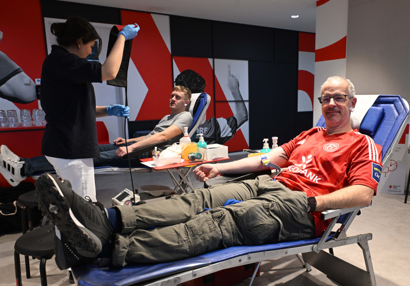 Zwei Männer liegen auf Liegen und spenden Blut. Eine Frau in schwarzem T-Shirt und blauen Handschuhen entnimmt einem der Männer Blut. Der Raum ist mit roten und schwarzen Grafiken an der Wand gestaltet.