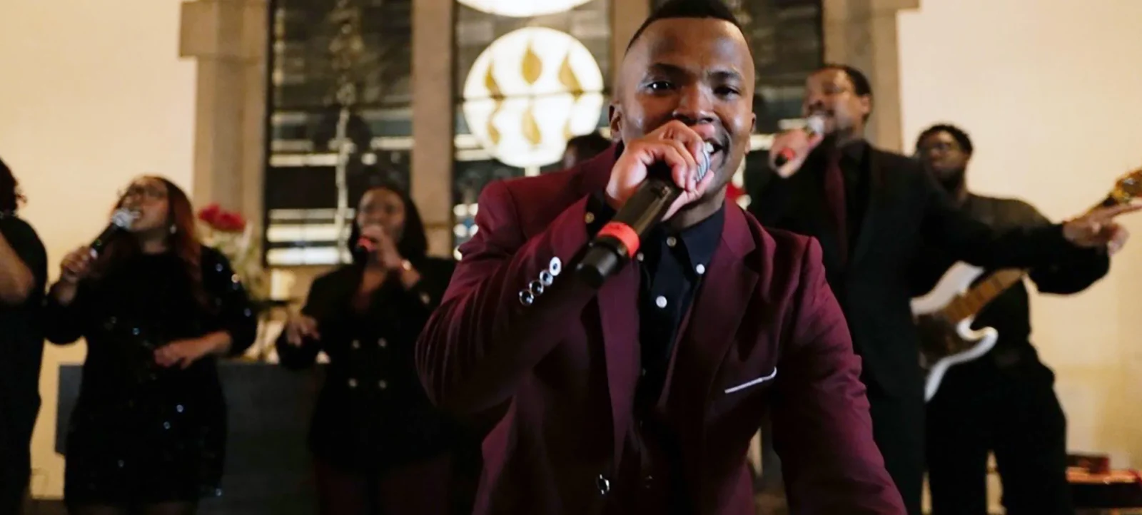 Eine Gruppe von Menschen singt gemeinsam in einem Raum mit großen Fenstern im Hintergrund. Ein Mann im Vordergrund hält ein Mikrofon und trägt einen roten Blazer. Die Atmosphäre wirkt festlich und harmonisch.