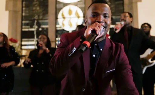 Eine Gruppe von Menschen singt gemeinsam in einem Raum mit großen Fenstern im Hintergrund. Ein Mann im Vordergrund hält ein Mikrofon und trägt einen roten Blazer. Die Atmosphäre wirkt festlich und harmonisch.