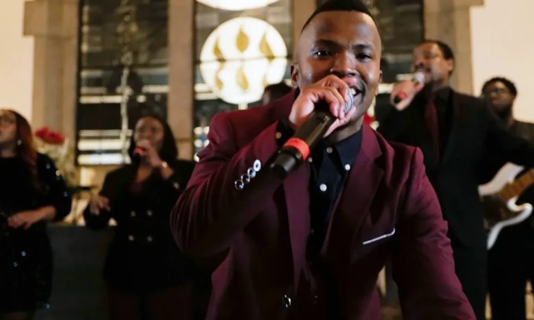 Eine Gruppe von Menschen singt gemeinsam in einem Raum mit großen Fenstern im Hintergrund. Ein Mann im Vordergrund hält ein Mikrofon und trägt einen roten Blazer. Die Atmosphäre wirkt festlich und harmonisch.