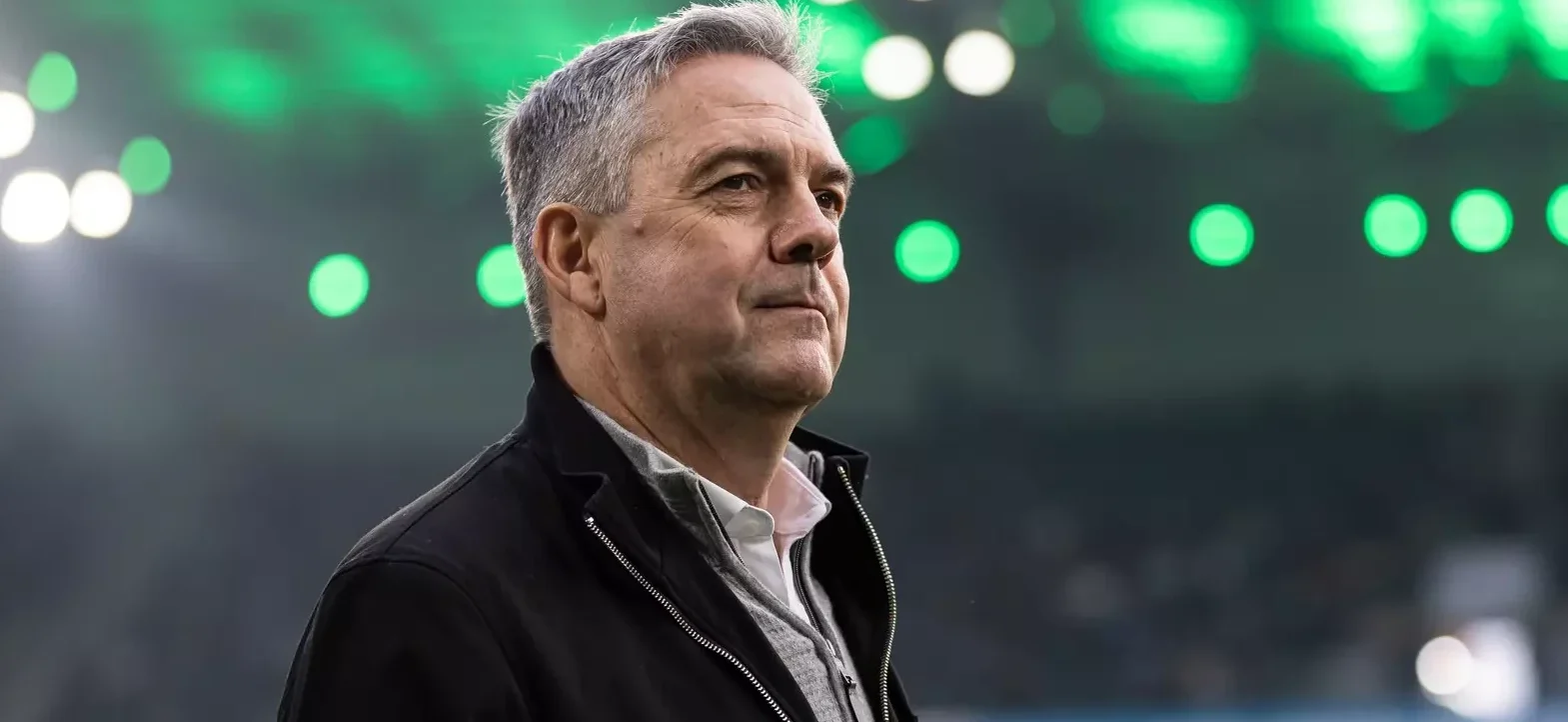 Ein Mann mit grauem Haar und einem schwarzen Jackett steht auf einem Fußballfeld und blickt in die Ferne. Im Hintergrund sind unscharfe grüne und weiße Lichter zu sehen, was auf eine Stadionatmosphäre hindeutet.