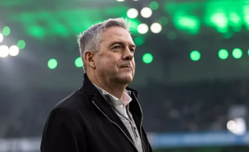 Ein Mann mit grauem Haar und einem schwarzen Jackett steht auf einem Fußballfeld und blickt in die Ferne. Im Hintergrund sind unscharfe grüne und weiße Lichter zu sehen, was auf eine Stadionatmosphäre hindeutet.