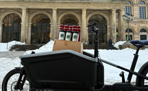 Ein Lastenfahrrad mit der Aufschrift "CUBE" steht vor einem historischen Gebäude mit großen Säulen und Bogenfenstern. Auf dem Fahrrad sind mehrere Getränkedosen und eine Pappkiste mit dem Logo "96" geladen. Der Boden ist mit Schnee bedeckt, und im Hintergrund ist eine Person zu sehen, die das Gebäude betritt.