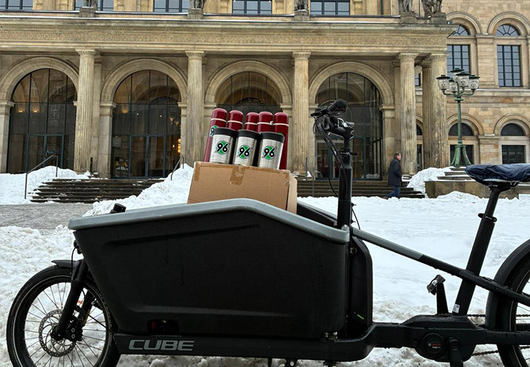 Ein Lastenfahrrad mit der Aufschrift "CUBE" steht vor einem historischen Gebäude mit großen Säulen und Bogenfenstern. Auf dem Fahrrad sind mehrere Getränkedosen und eine Pappkiste mit dem Logo "96" geladen. Der Boden ist mit Schnee bedeckt, und im Hintergrund ist eine Person zu sehen, die das Gebäude betritt.