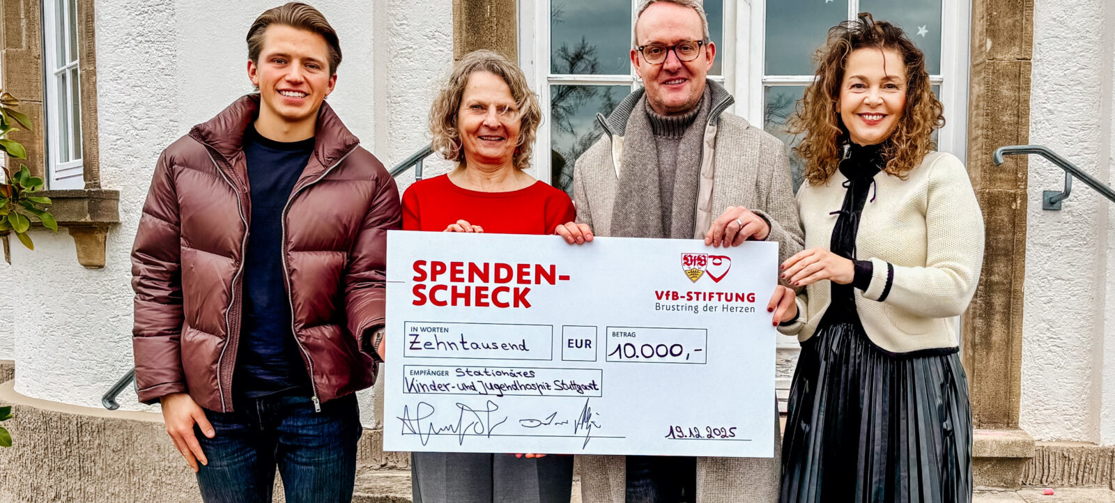 Vier Personen stehen vor einem Gebäude und halten einen großen Scheck der VfB-Stiftung in Höhe von 10.000 Euro. Der Scheck ist für die Stationäres Kinder- und Jugendhospiz Stuttgart bestimmt. Die Personen lächeln und wirken zufrieden.