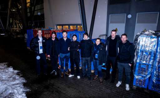 Eine Gruppe von Menschen steht vor einem Gebäude und hält blaue Säcke mit Essensspenden. Sie lächeln in die Kamera. Im Hintergrund sind weitere Säcke und ein schneebedeckter Boden zu sehen.
