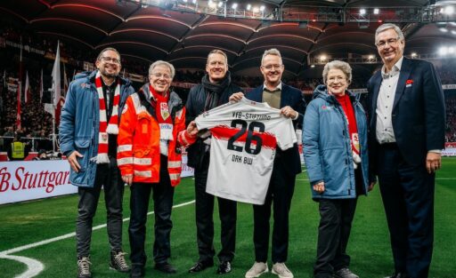 Eine Gruppe von fünf Personen steht auf dem Rasen eines Fußballstadions und hält ein Trikot des VfB Stuttgart mit der Aufschrift "VfB-Stiftung" und der Nummer 26 hoch. Im Hintergrund sind Tribünen mit Zuschauern und Werbebanner zu sehen. Die Personen lächeln und wirken zufrieden, was auf eine erfolgreiche oder bedeutende Veranstaltung hindeutet.