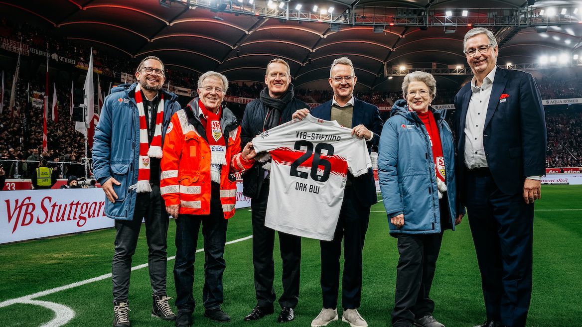 Eine Gruppe von fünf Personen steht auf dem Rasen eines Fußballstadions und hält ein Trikot des VfB Stuttgart mit der Aufschrift "VfB-Stiftung" und der Nummer 26 hoch. Im Hintergrund sind Tribünen mit Zuschauern und Werbebanner zu sehen. Die Personen lächeln und wirken zufrieden, was auf eine erfolgreiche oder bedeutende Veranstaltung hindeutet.