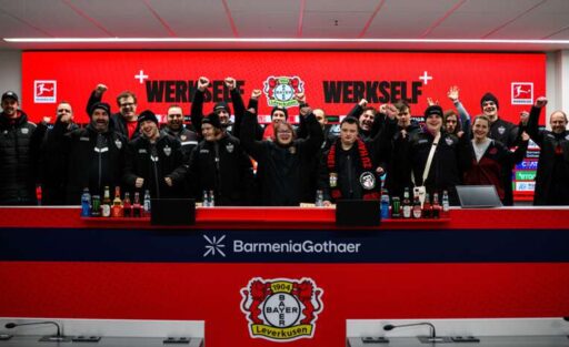 Gruppe von Menschen bei einer Pressekonferenz des Fußballvereins Bayer 04 Leverkusen, mit dem Trainer Kasper Hjulmand in der Mitte. Die Teilnehmer halten ein Schild mit der Aufschrift "Werkself" und dem Vereinslogo. Im Hintergrund ist das Logo des Sponsors BarmeniaGothaer zu sehen.