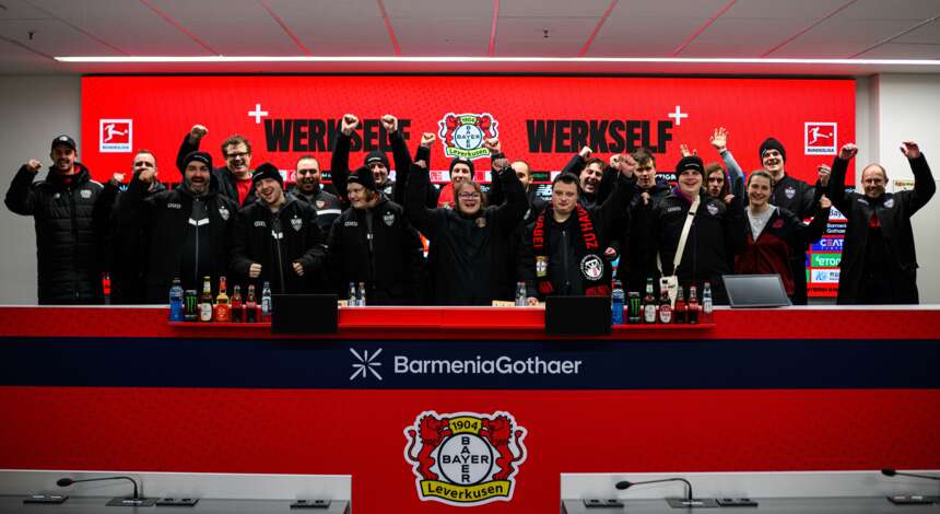 Gruppe von Menschen bei einer Pressekonferenz des Fußballvereins Bayer 04 Leverkusen, mit dem Trainer Kasper Hjulmand in der Mitte. Die Teilnehmer halten ein Schild mit der Aufschrift "Werkself" und dem Vereinslogo. Im Hintergrund ist das Logo des Sponsors BarmeniaGothaer zu sehen.