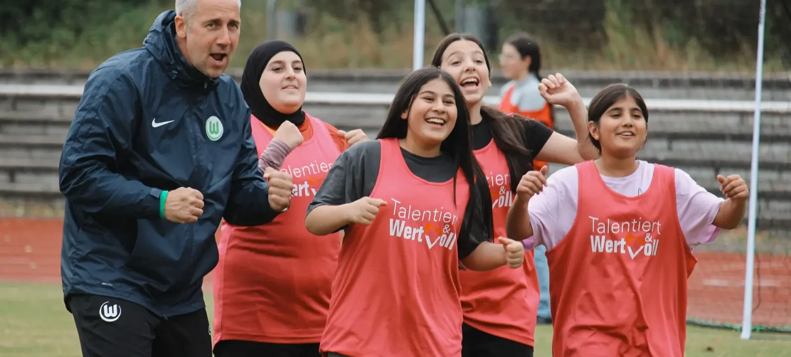 Eine Gruppe von Menschen, darunter ein Mann und vier Frauen, stehen auf einem Fußballfeld und lächeln in die Kamera. Der Mann trägt eine dunkle Jacke mit einem grünen Logo, während die Frauen rote Trainingswesten mit der Aufschrift "Talentiert & Wertvoll" tragen. Im Hintergrund sind Tribünen und ein Fußballtor zu sehen.