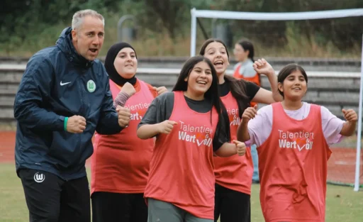 Eine Gruppe von Menschen, darunter ein Mann und vier Frauen, stehen auf einem Fußballfeld und lächeln in die Kamera. Der Mann trägt eine dunkle Jacke mit einem grünen Logo, während die Frauen rote Trainingswesten mit der Aufschrift "Talentiert & Wertvoll" tragen. Im Hintergrund sind Tribünen und ein Fußballtor zu sehen.