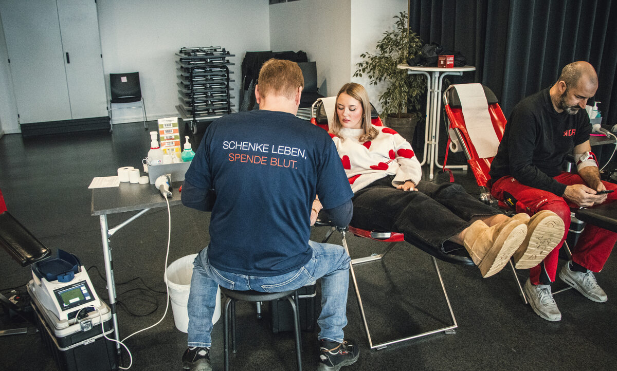 Drei Personen bei einer Blutspendeaktion. Eine Person sitzt auf einem Stuhl und spendet Blut, während eine andere Person, die ein T-Shirt mit der Aufschrift "Schenke Leben, spende Blut" trägt, die Blutentnahme durchführt. Eine dritte Person sitzt ebenfalls auf einem Stuhl und spendet Blut. Im Hintergrund sind Stapel von Klappstühlen und ein Tisch mit Desinfektionsmitteln zu sehen.