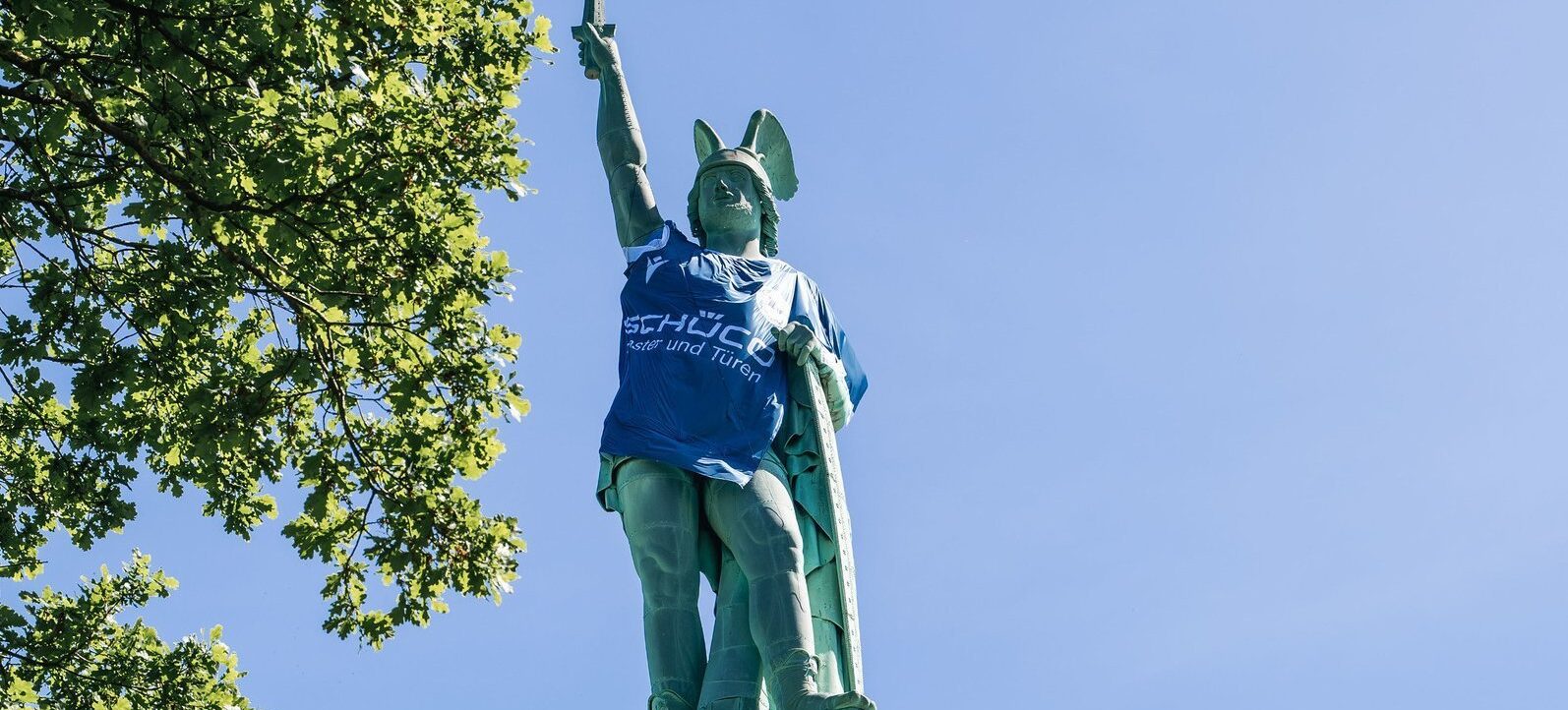 Eine große Statue eines Kriegers mit Helm und Schild, der eine Fahne in die Höhe hält. Die Statue trägt ein blaues T-Shirt mit der Aufschrift 'SCHÜCO Meister und Pokalsieger'. Die Statue steht auf einem hohen Sockel und ist von grünen Bäumen umgeben, bei klarem Himmel.