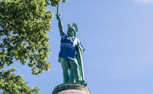 Eine große Statue eines Kriegers mit Helm und Schild, der eine Fahne in die Höhe hält. Die Statue trägt ein blaues T-Shirt mit der Aufschrift 'SCHÜCO Meister und Pokalsieger'. Die Statue steht auf einem hohen Sockel und ist von grünen Bäumen umgeben, bei klarem Himmel.