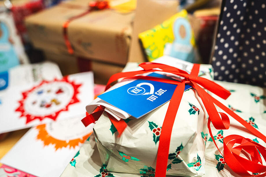 Ein Geschenk mit weißem Geschenkpapier, das mit grünen Stechpalmen und roten Bändern verziert ist. Auf dem Geschenk befindet sich ein blaues Etikett mit dem Logo der DFL und der Aufschrift "Tut Gutes". Im Hintergrund sind weitere Geschenke und Weihnachtskarten zu sehen.