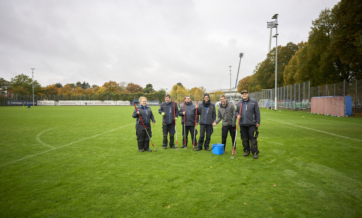 Eine Gruppe von sechs Personen steht auf einem Fußballfeld und hält jeweils eine Vertikutierwalze in den Händen. Sie tragen dunkle Arbeitskleidung und stehen auf einem gut gepflegten Rasen. Im Hintergrund sind Werbebanner und ein Zaun zu sehen.